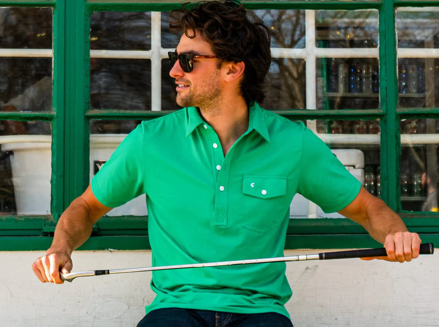 Man wearing Criquet Performance Jersey Players Shirt in Augusta Green, holding a golf club in front of a window.
